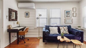 A living room with a dark blue sofa, a wooden desk, a chair, and a floor lamp. There are framed pictures on the wall and a window with blinds. - Remodeled Designer Cottage with Bicycles (Culver City)