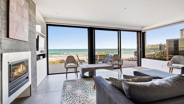 A modern living room with a fireplace, seating area, and large glass doors leading to a beach view. - Aqua Mist Seascape - refreshing ocean-views (Port Fairy)