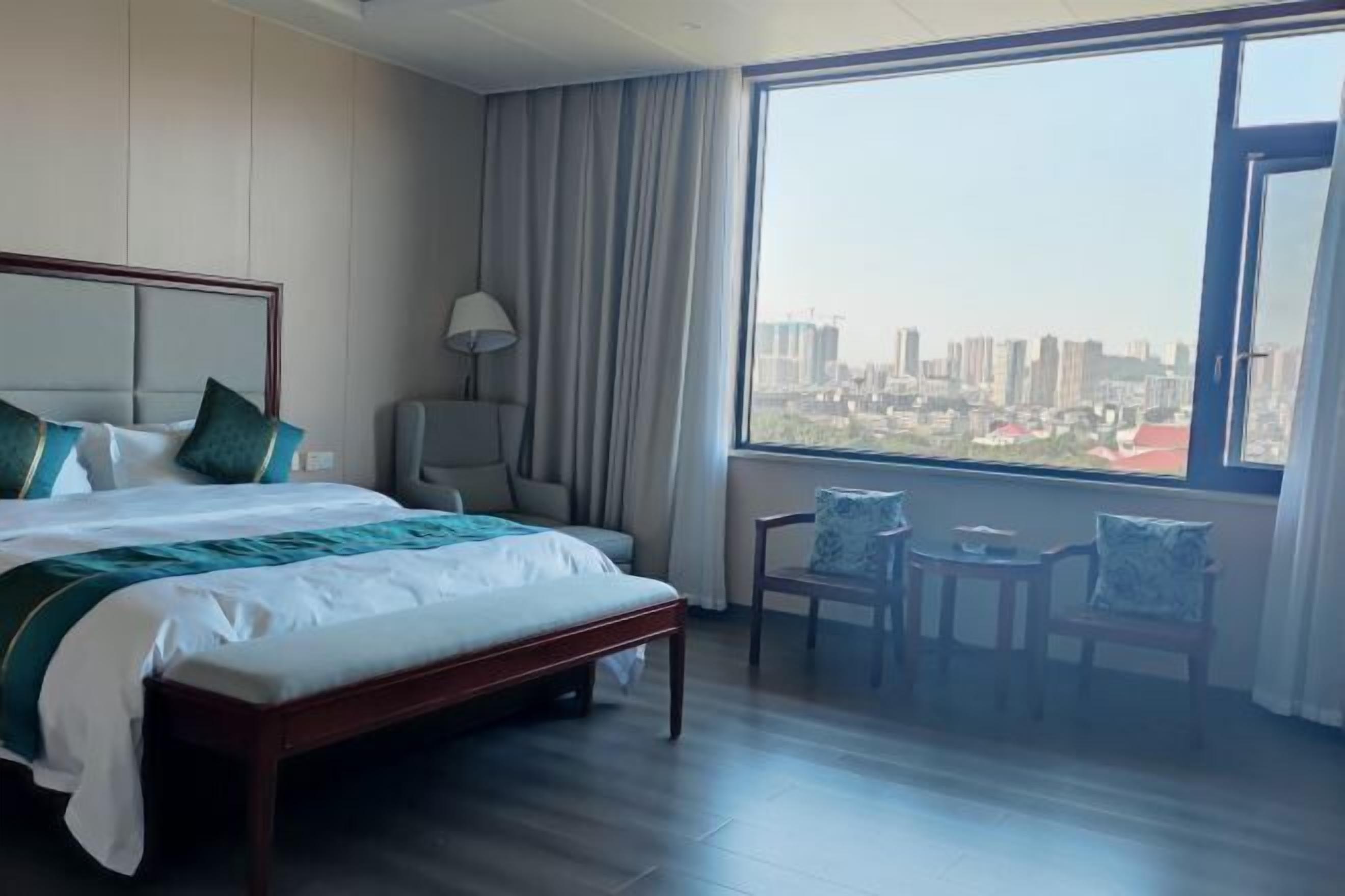 A hotel room with a large bed, a desk, two chairs, and a view of a cityscape.