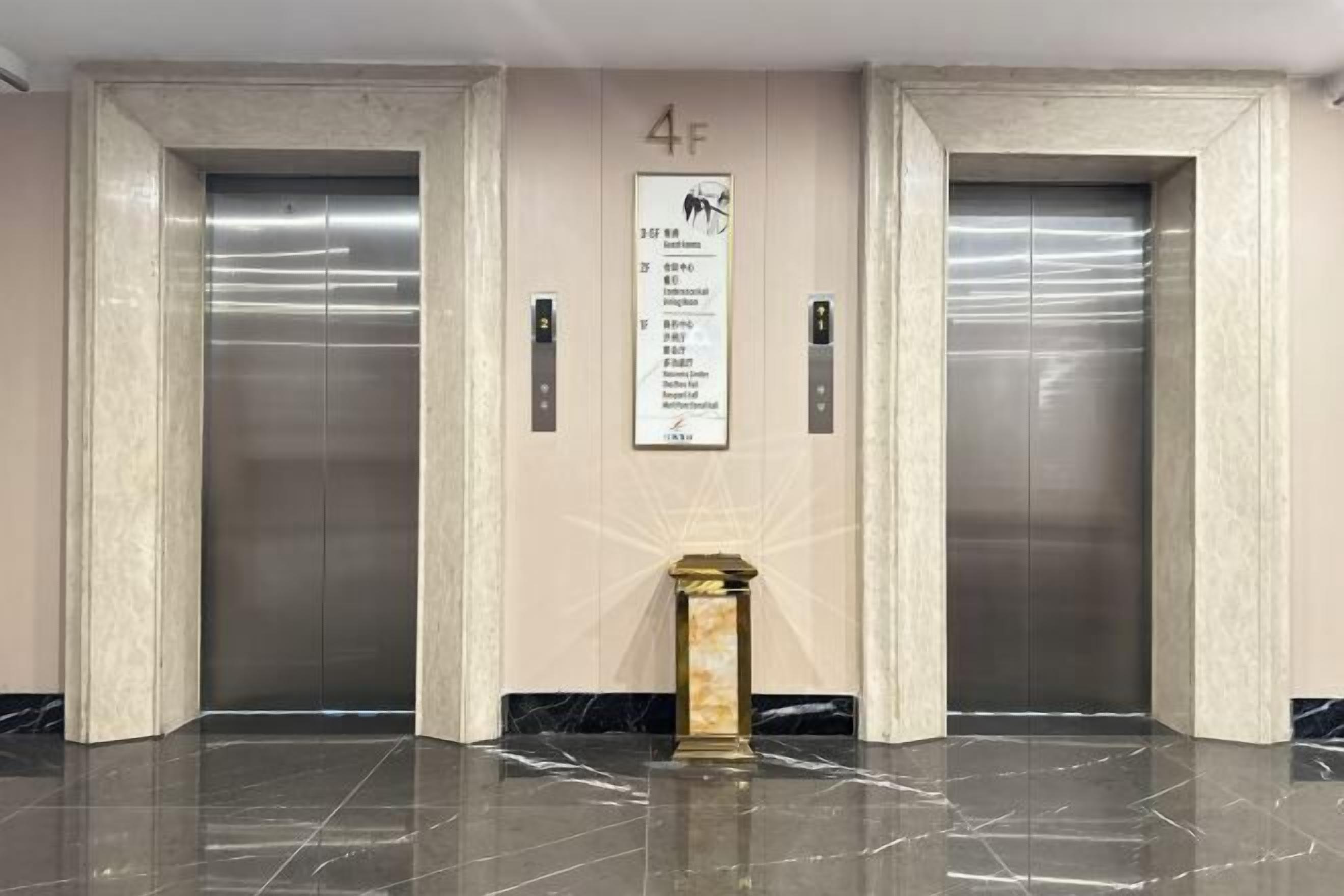 Un espace comprenant deux ascenseurs, un sol en marbre et un panneau mural indiquant les consignes de sécurité en cas d’urgence.