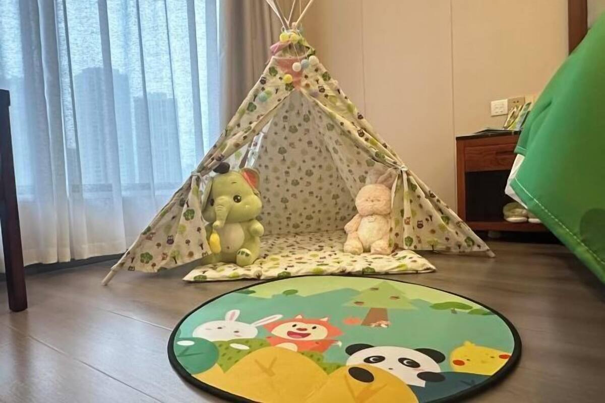 A room with a play tent, a round rug with cartoon characters, a green couch, and a window with curtains.