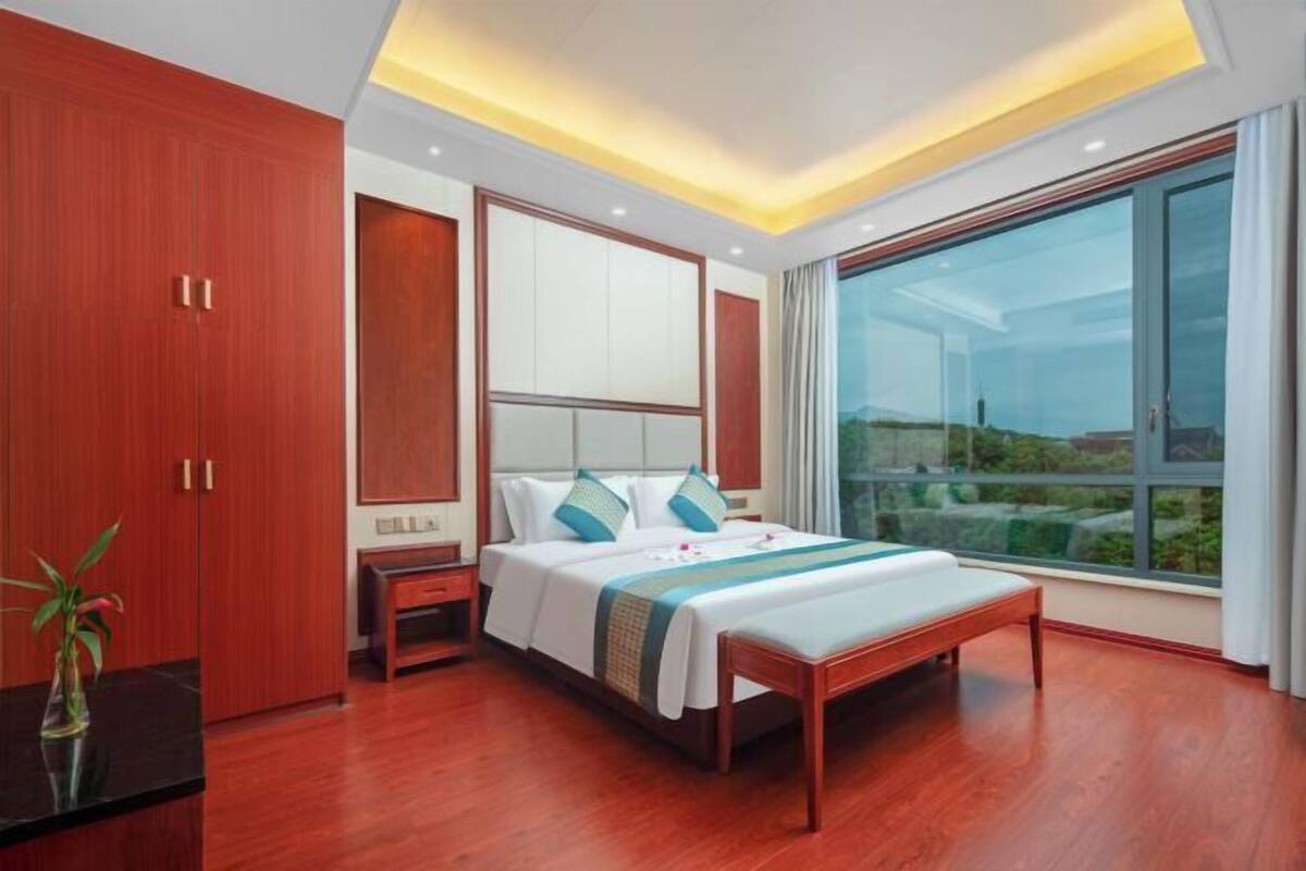 A modern hotel room with a large bed, wooden wardrobe, bedside table, and a large window offering a view.