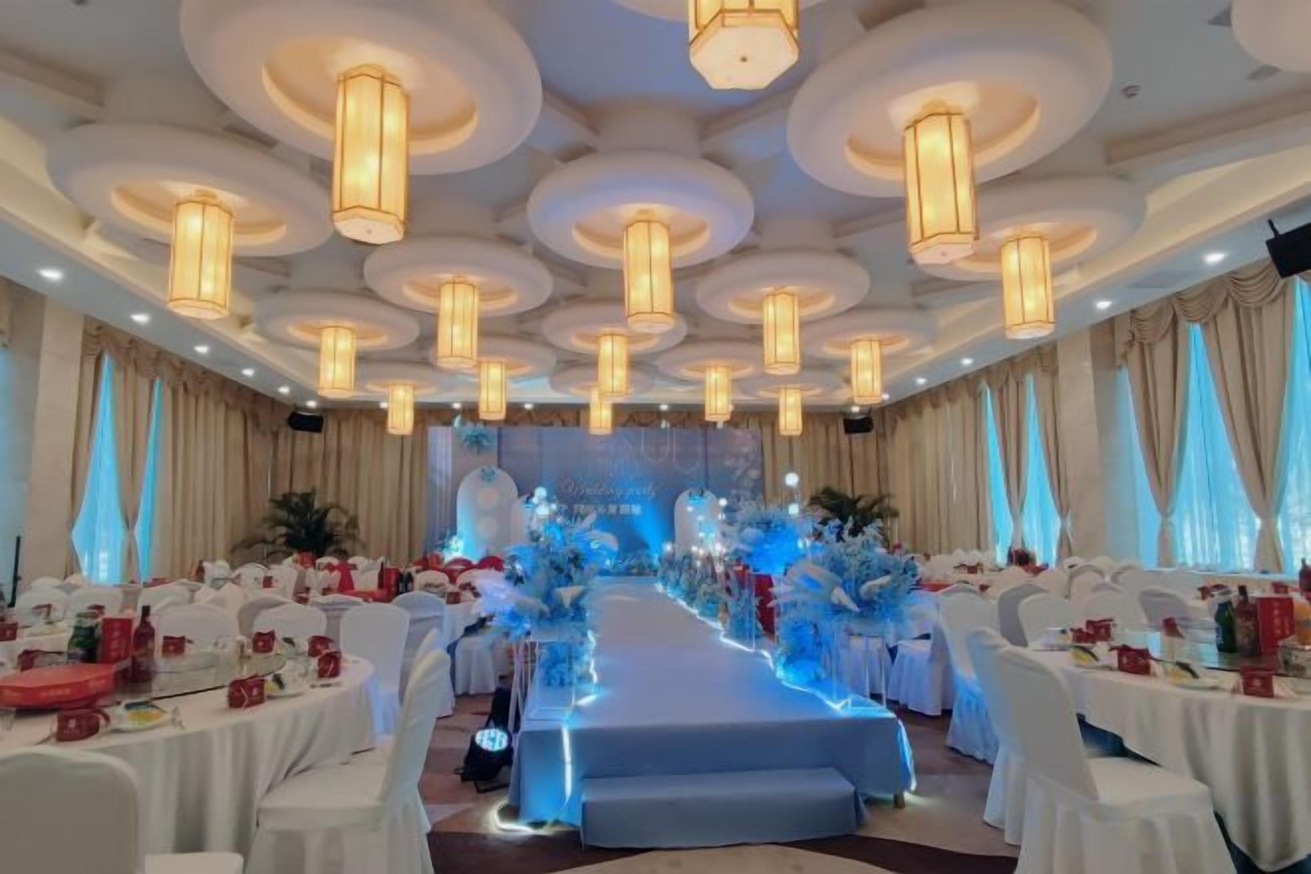 Une salle de banquet avec des tables rondes, une scène décorée de fleurs et des lustres.