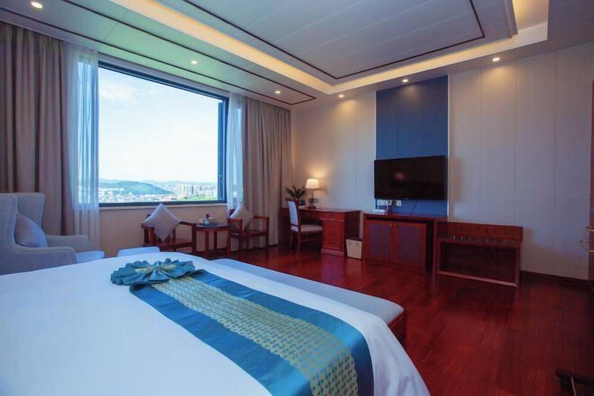 A hotel room with a large bed, a desk, a chair, a TV, and a window with a city view.