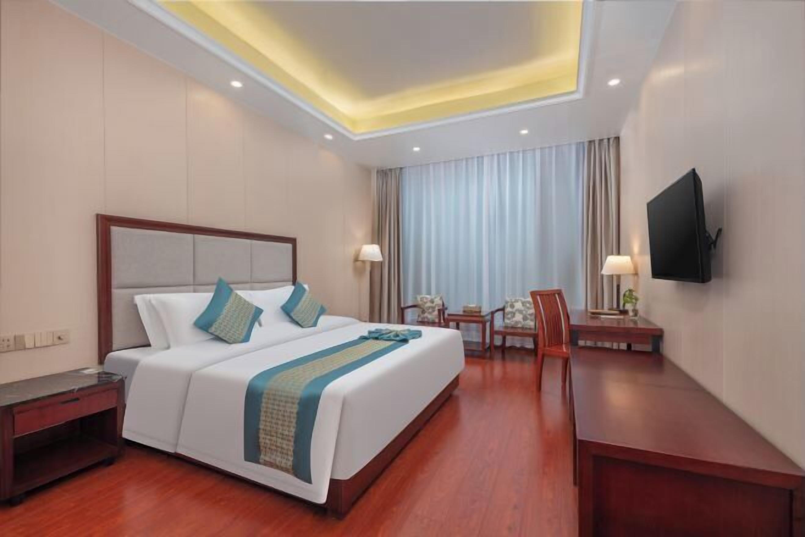 A hotel room with a large bed, bedside tables, a desk, a chair, a TV, and a window with curtains.