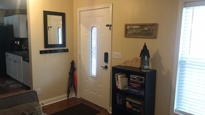 A living room with a sofa, a bookshelf with books and decorative items, a door with a window, and a kitchen area in the background.