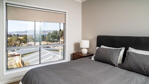 A bedroom with a large window, a bed with grey bedding, a nightstand with a lamp, and a view of a landscape with trees and buildings. - Aspect - 2/8a Poley Cow Lane (Jindabyne)