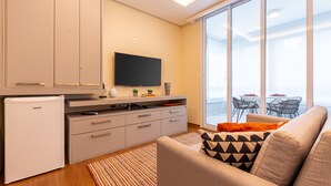 A modern living room with a sofa, a flat-screen TV, a mini-fridge, and a sliding glass door leading to a balcony with outdoor furniture. - 100m from the Avenue. Paulista / 30m from the shopping mall / close to the subway and hospitals (Bela Vista)