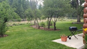Un patio trasero con césped, árboles, una parrilla de barbacoa y una terraza de madera.