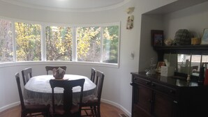 Un comedor con una mesa redonda y sillas, una ventana con vistas a árboles, y un mueble de madera oscura con varios objetos encima.