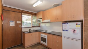 A compact kitchen with wooden cabinets, a microwave, a stove, and a refrigerator. There is a window with blinds, a sink, and a fire extinguisher.