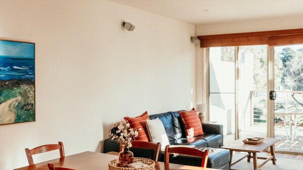 Sala de estar com mesa de jantar, cadeiras, sofá com almofadas, um quadro na parede e vista através de uma porta de vidro.