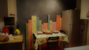 Una cuina amb una taula per a dues persones, un frigorífic i una paret amb panells de colors.