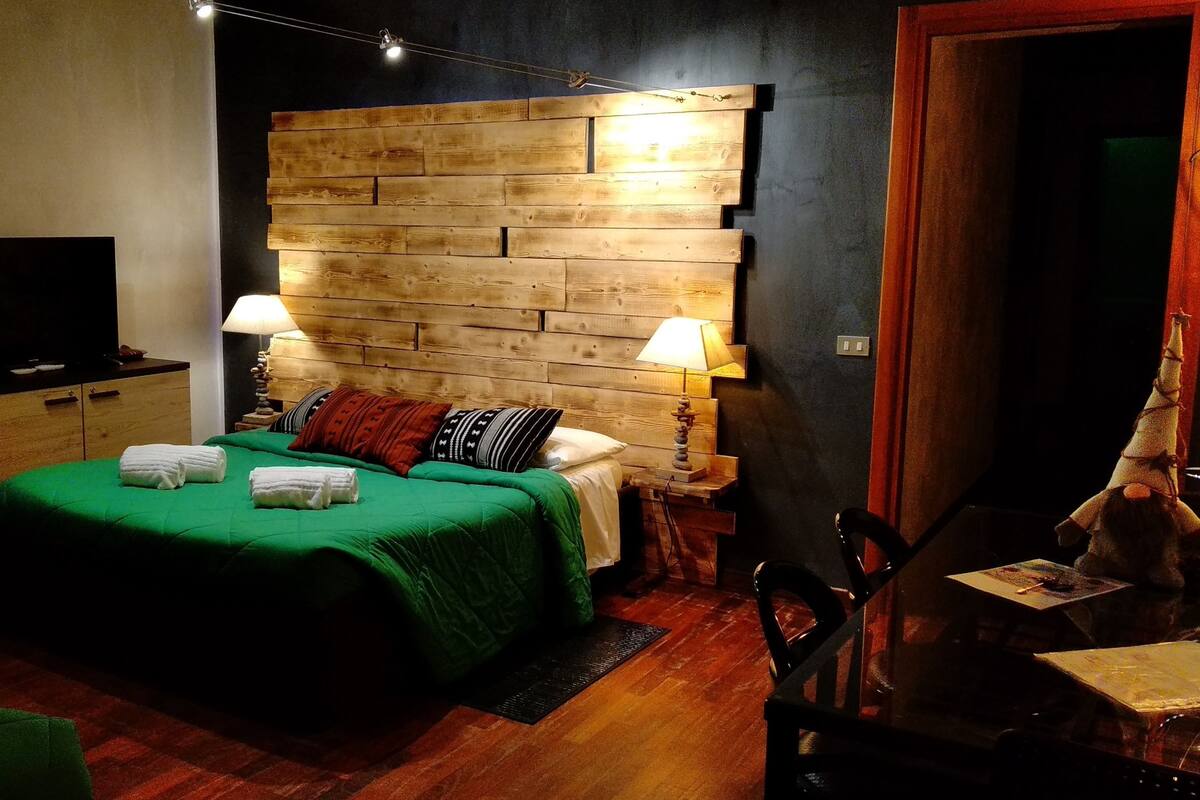 A hotel room with a wooden headboard, a bed with green bedding, two bedside lamps, a TV on a wooden cabinet, and a small table with a chair.