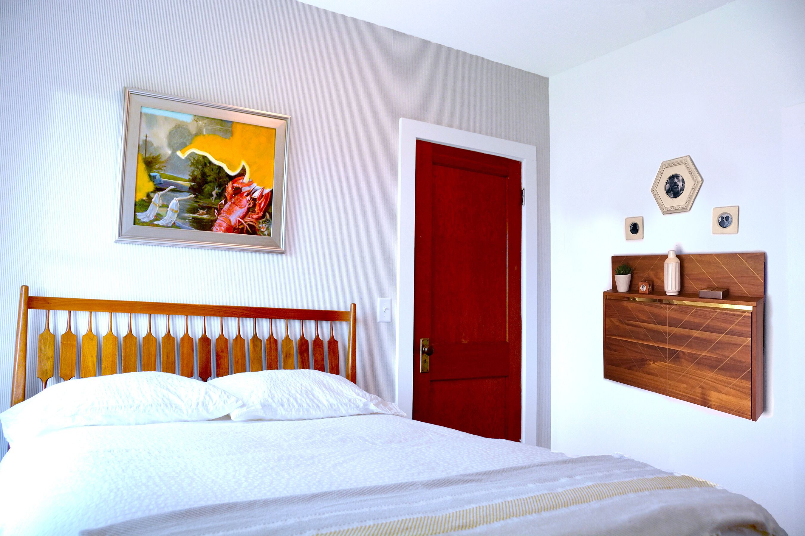A bedroom with a wooden headboard, a red door, a wall-mounted shelf with decorative items, and a framed painting above the bed.