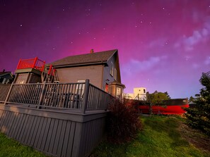 Une maison avec une terrasse en bois, une rambarde rouge et un ciel nocturne dégagé parsemé d’étoiles.