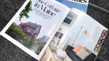 A magazine spread featuring a cottage in Maine with a glass table, a sofa, and a bookshelf.