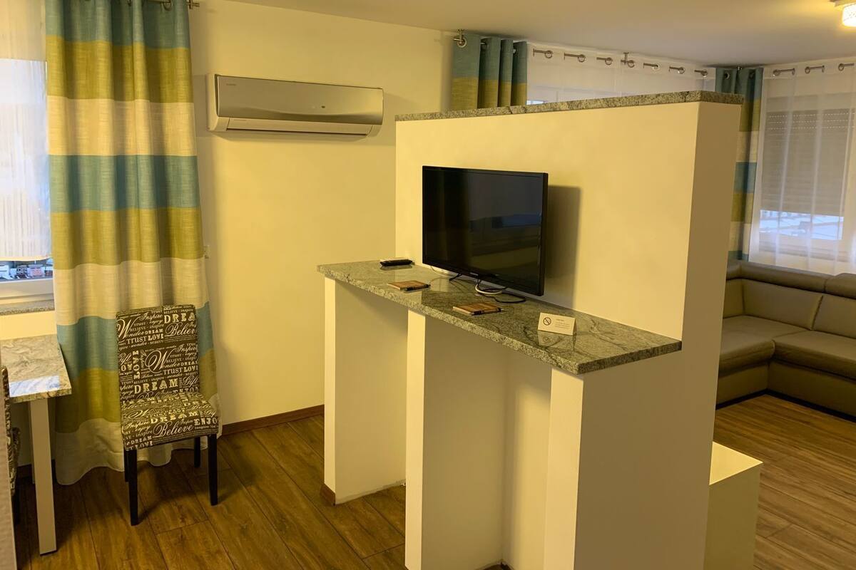 A compact room with a TV on a stand, a small table, a chair with a motivational quote cushion, a sofa, and an air conditioning unit.