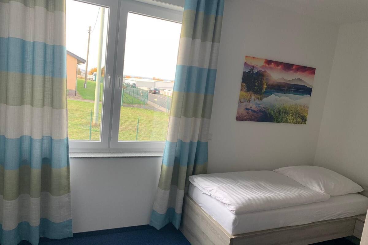 A single-bed room with a window, curtains, and a painting on the wall.