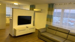 A modern living room with a flat-screen TV on a white entertainment unit, a beige sofa, and a large window with curtains. - Hotel Am Spielacker (Gelnhausen)