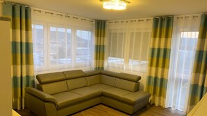 A living room with a sectional sofa, large windows with sheer curtains, and a wooden floor. - Hotel Am Spielacker (Gelnhausen)