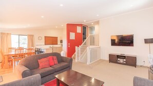 A living room with a sofa, a coffee table, a TV, a staircase, and a dining area with a table and chairs. - Boronia - 5/30 Nettin Circuit (Jindabyne)
