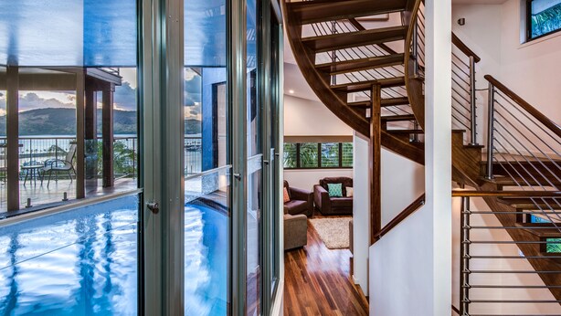 Un interior moderno con un ventanal de vidrio que da a una piscina, una escalera de madera y un área de estar con vistas al exterior.