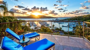 Un balcón con sillones reclinables azules y una mesa, con vistas a un puerto deportivo con barcos y la vista del atardecer.