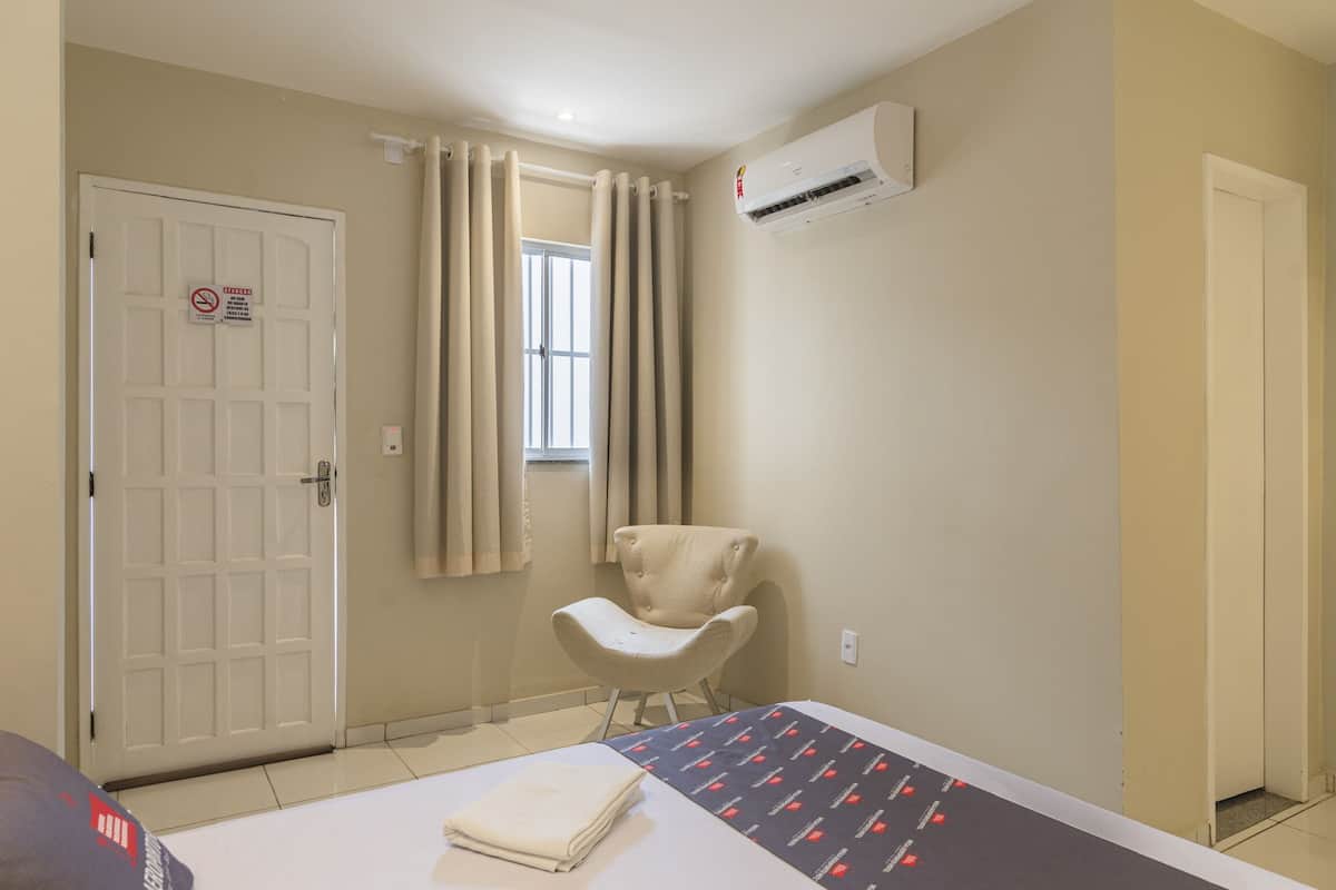 A hotel room with a bed, a chair, a window with curtains, an air conditioning unit, and a door with a sign.