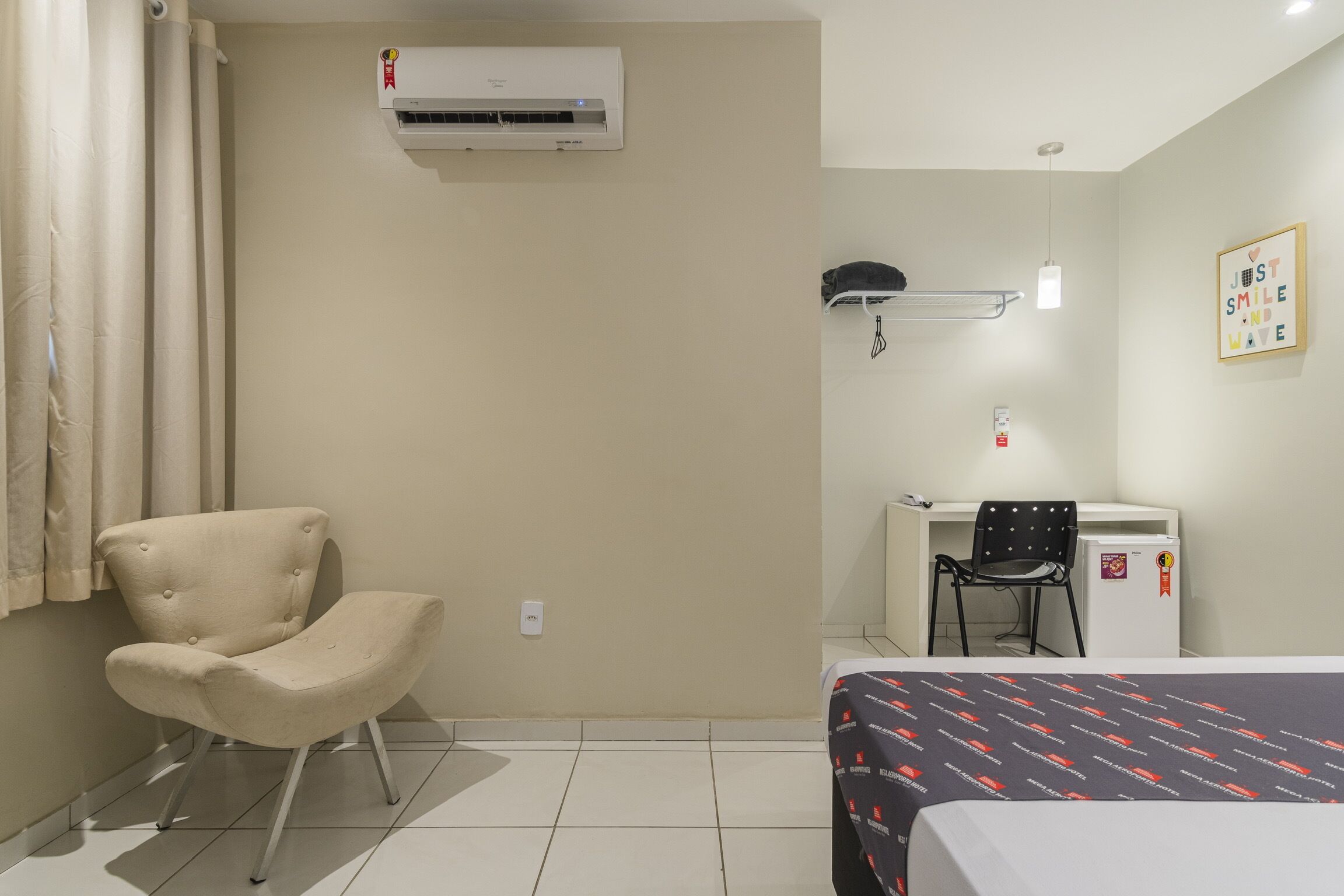 Una habitación de hotel con cama, una silla, escritorio, unidad de aire acondicionado y un refrigerador pequeño.