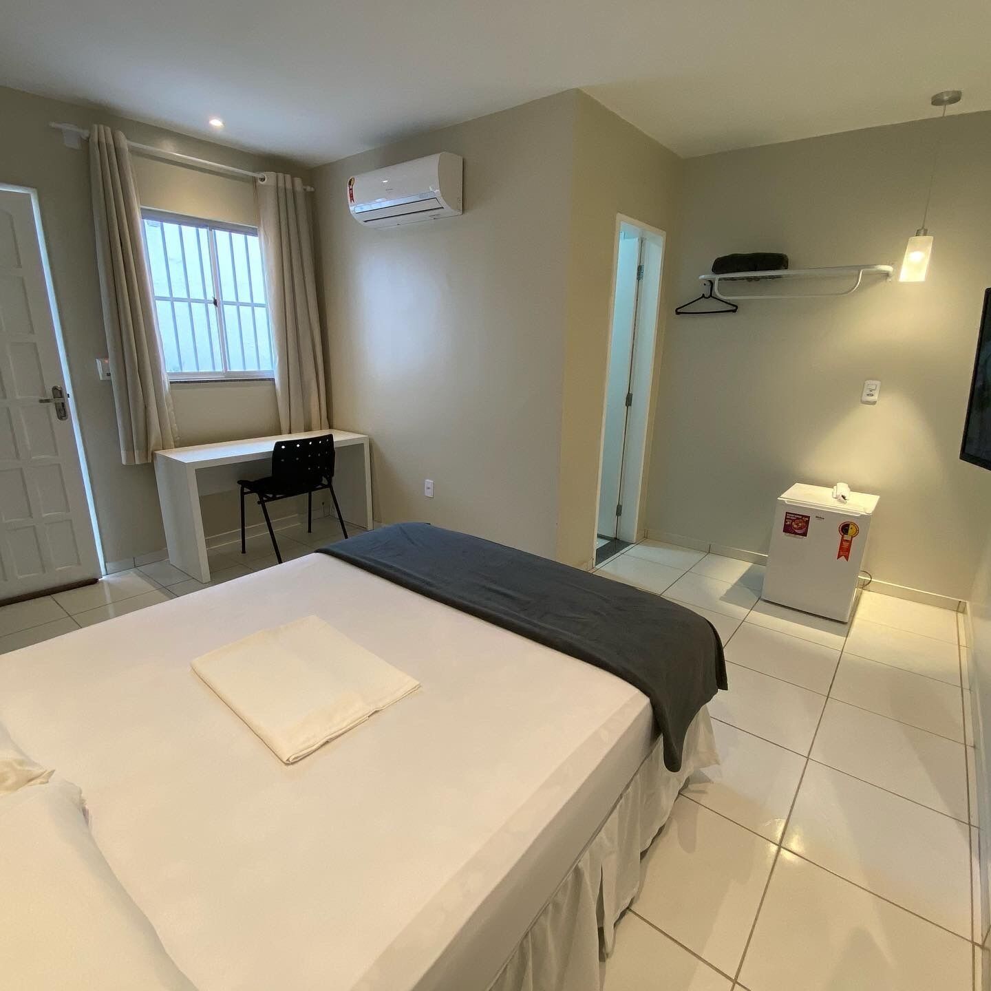 A hotel room with a bed, a desk, a chair, a wardrobe, a mirror, a TV, and a small refrigerator.