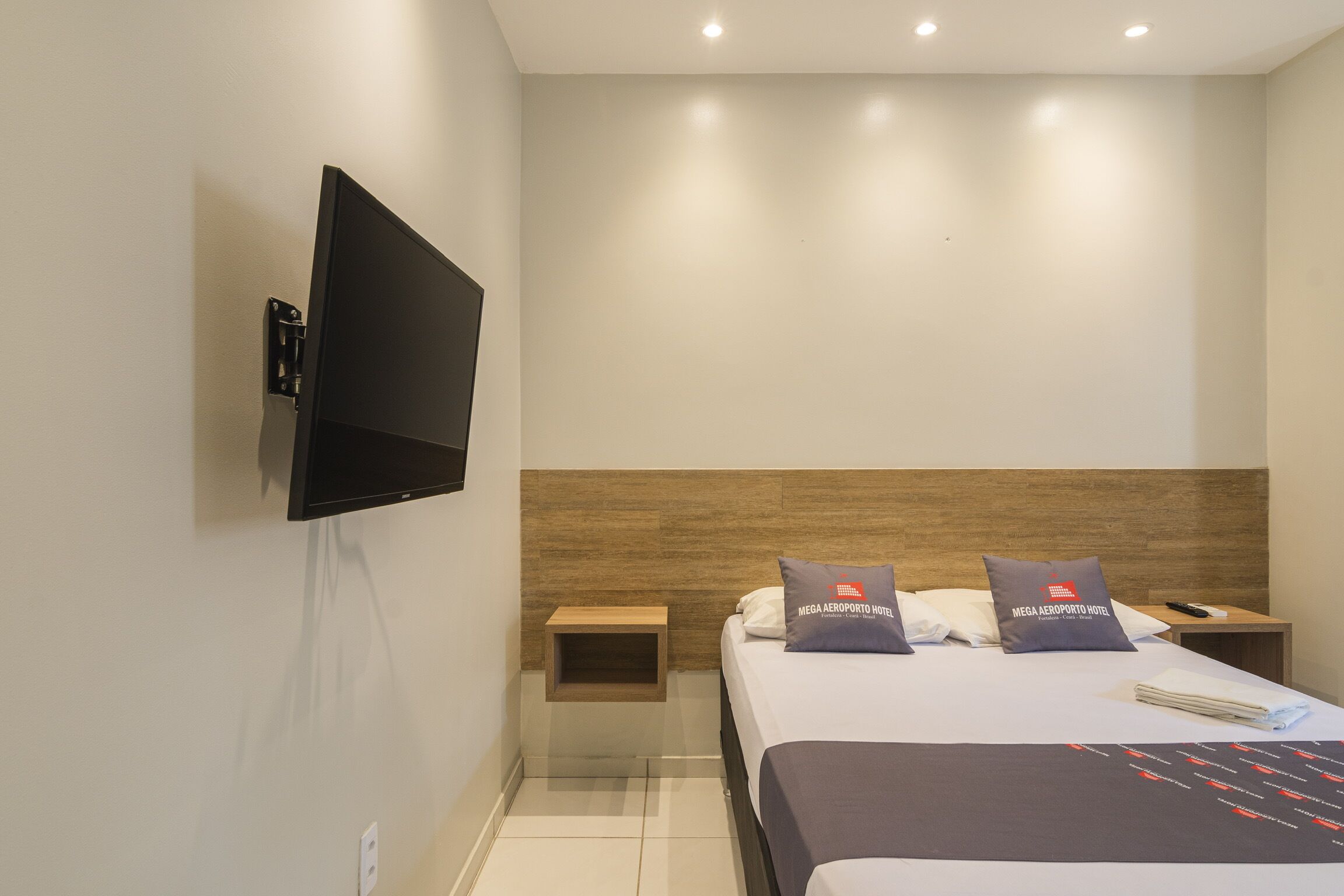 A modern hotel room with a flat-screen TV mounted on the wall, a wooden headboard, two pillows with the hotel's logo, a bedside table with a remote control, and a neatly made bed.