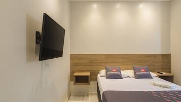 A modern hotel room with a flat-screen TV mounted on the wall, a wooden headboard, two pillows with the hotel's logo, a bedside table with a remote control, and a neatly made bed.