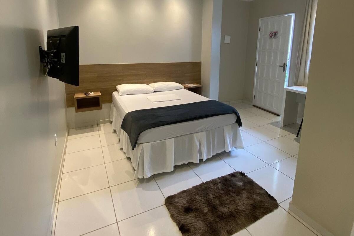 A bedroom with a bed, a TV mounted on the wall, a door, and a small wooden shelf.