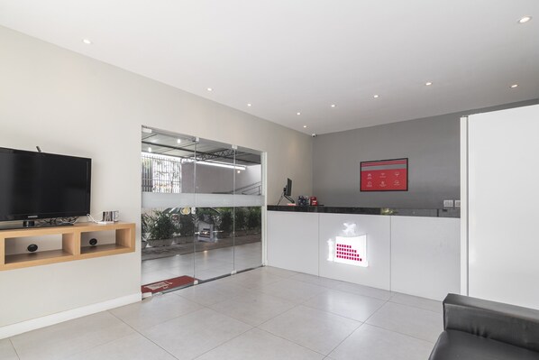 A modern living room with a flat-screen TV, wooden shelves, a glass door leading to a balcony, and a red emergency exit sign. - Mega Aeroporto Hotel (Fortaleza)