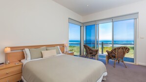 A bedroom with a large bed, a nightstand, two wicker chairs, and a view of the ocean through floor-to-ceiling windows. - Wytonia 1 - Spectacular Ocean Views (Port Fairy)