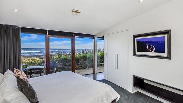 A bedroom with a large bed, a TV mounted on the wall, and a sliding glass door leading to a balcony with a view of the ocean. - Hearns Beachside Villa 3 (Port Fairy)