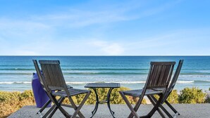 Dos sillones de madera y una pequeña mesa de metal sobre una superficie de concreto, con vista a una playa, el océano y un cielo despejado.