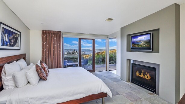 A bedroom with a large bed, a fireplace, a TV, and a balcony with a view of the ocean. - Hearns Beachside Villa 4 (Port Fairy)