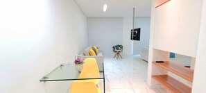 A modern living room with a glass coffee table, a yellow sofa, a white sofa with yellow cushions, a small side table with a vase of flowers, and a black TV on a stand. - Flats no Muro Alto Club (Ipojuca)