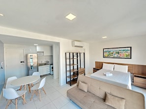 Quarto moderno e bem iluminado com cozinha compacta, área de jantar com mesa e cadeiras, sofá e cama com cabeceira.