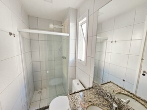 A bathroom with white tiled walls, a glass shower enclosure, a granite countertop, and a toilet. - Flats no Muro Alto Club (Ipojuca)