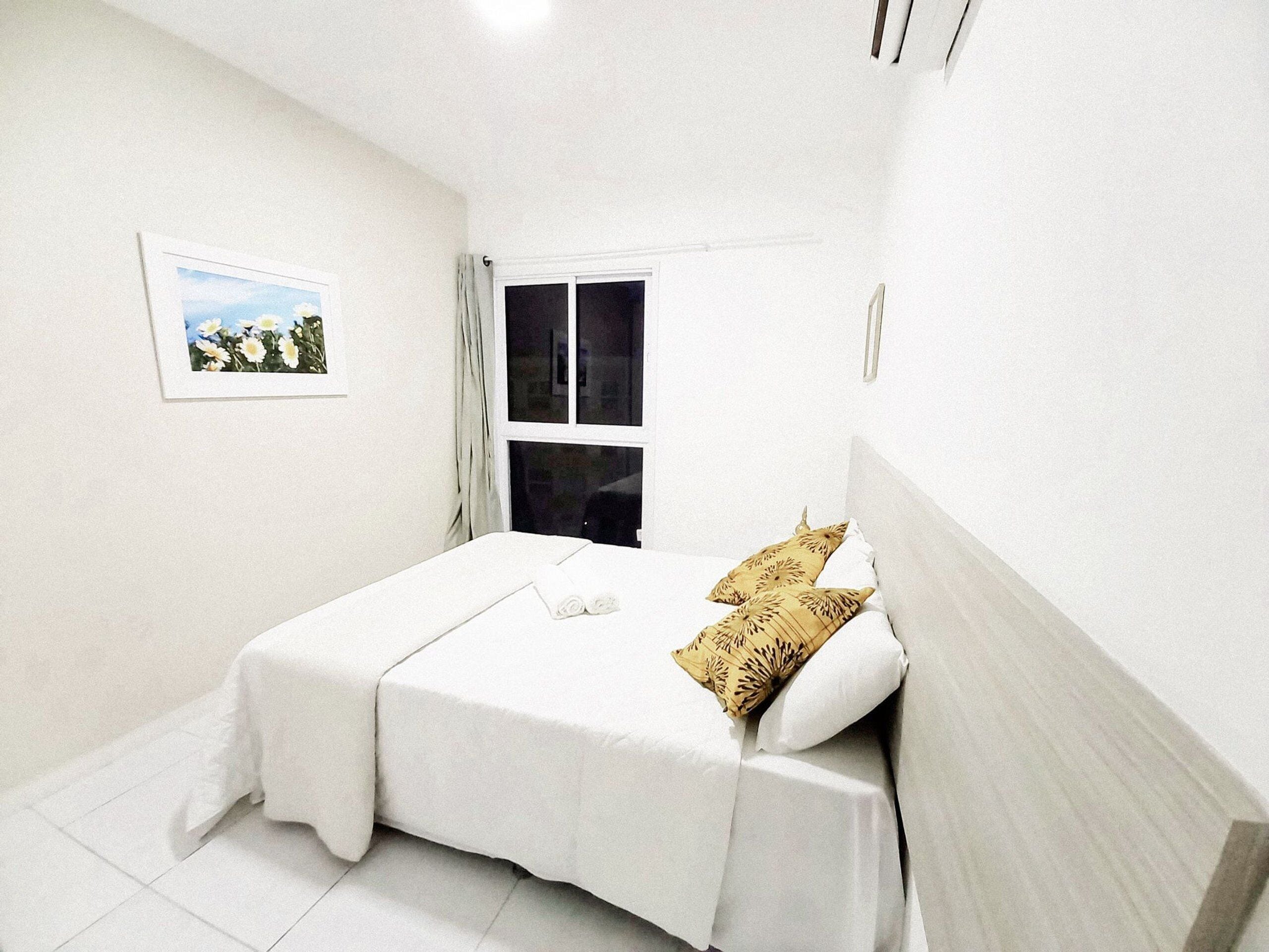 Un lit bien fait, avec une literie blanche et deux coussins décoratifs, une fenêtre avec des rideaux et un tableau encadré accroché au mur.