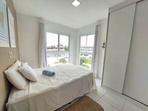 Quarto com cama, duas janelas, vista para uma piscina e edifícios, e um guarda-roupa.