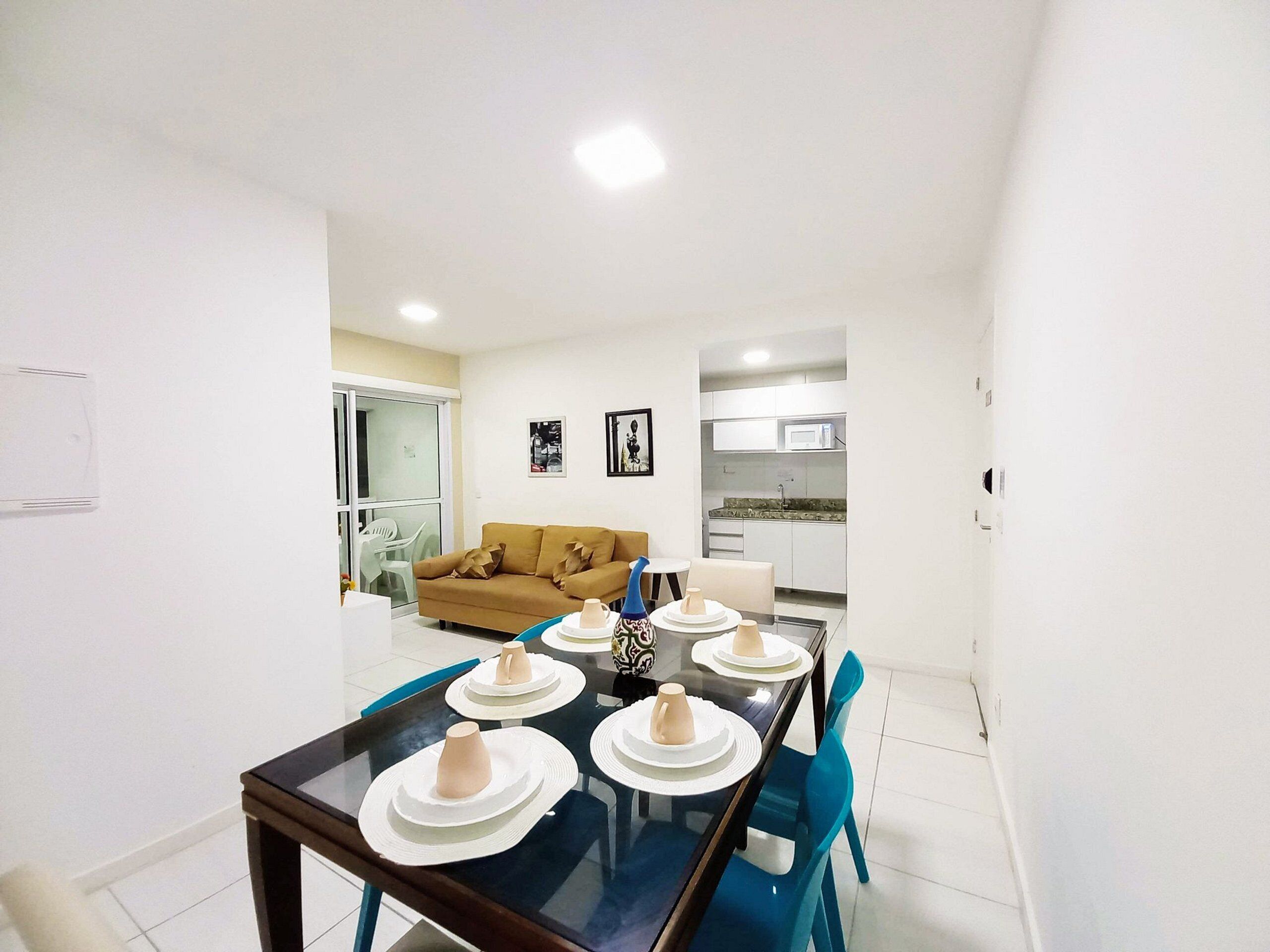 Un espacio de estar moderno con comedor que incluye una mesa de vidrio, sillas azules y platos blancos. Se ve un sofá y una cocina con armarios blancos al fondo.