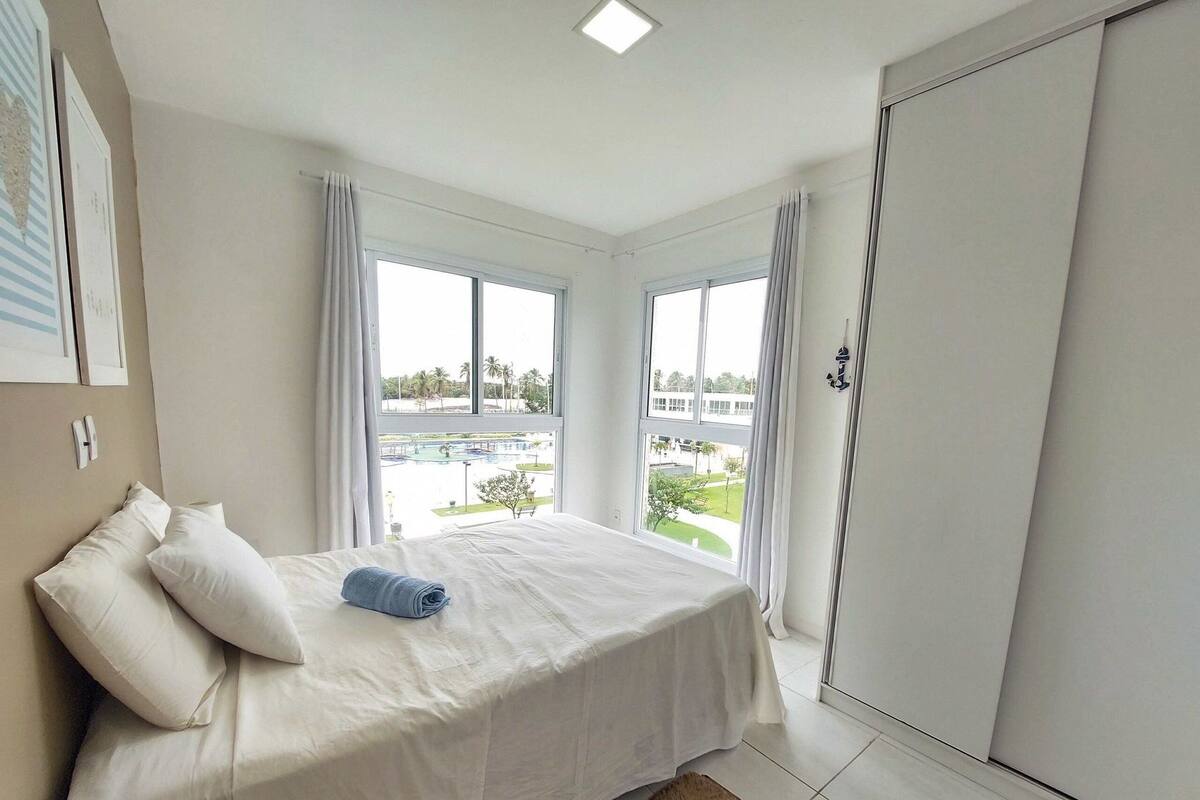 A bedroom with a bed, two windows, a view of a pool and buildings, and a wardrobe.