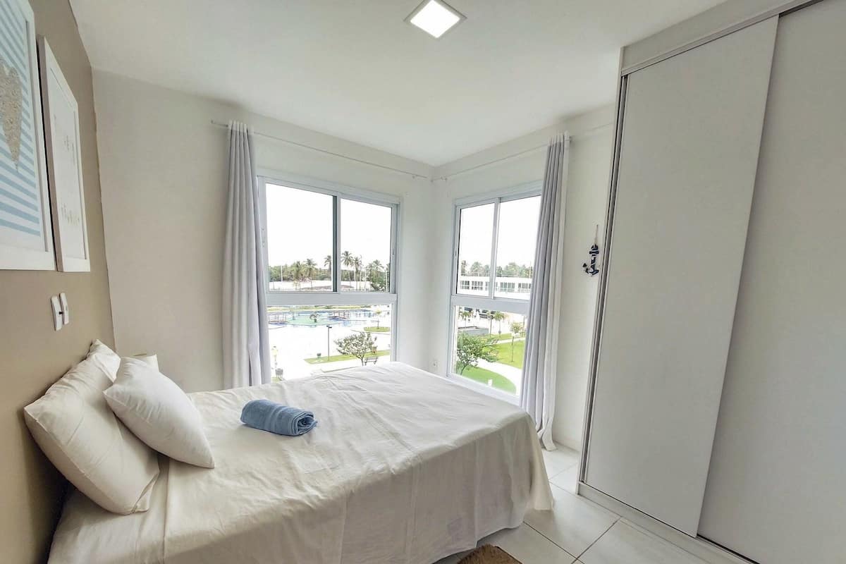 A bedroom with a bed, two windows, a view of a pool and buildings, and a wardrobe.