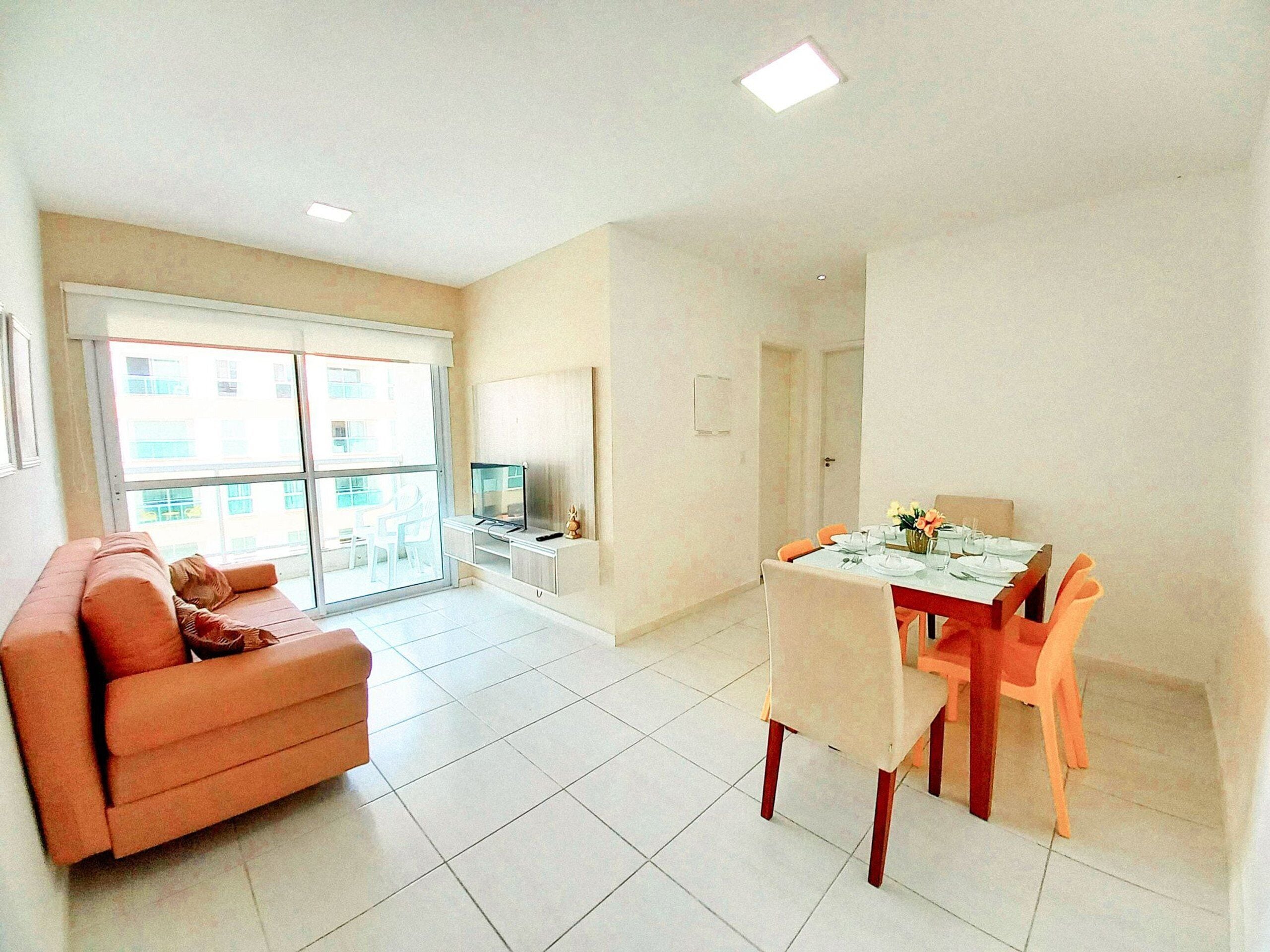 Uma sala de estar com sofá, mesa de jantar com cadeiras, televisão e varanda com vista para edifícios.