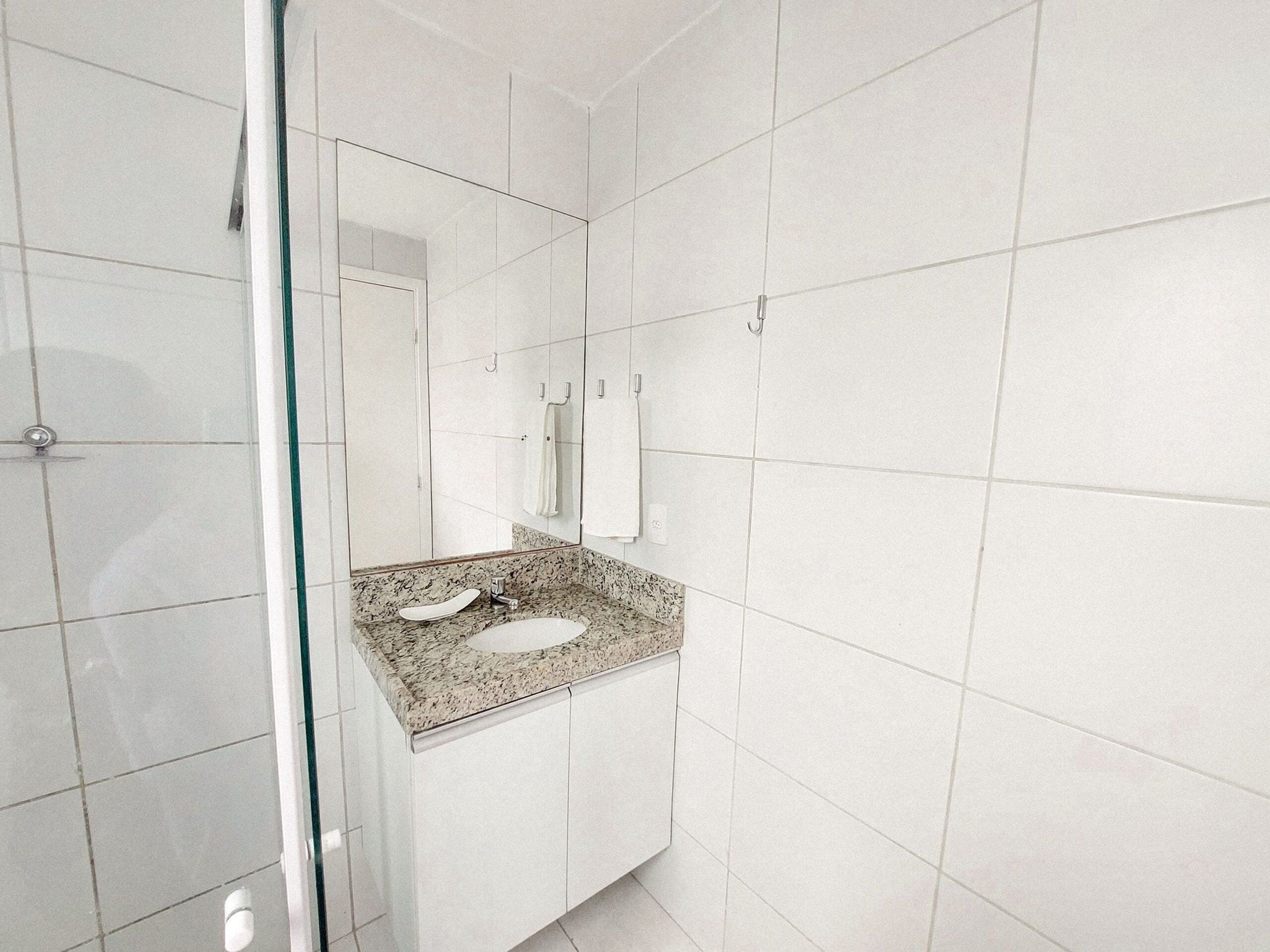 A modern bathroom with white tiles, a granite countertop sink, a glass shower enclosure, and a large mirror.