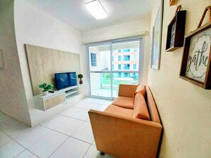 A modern living room with a flat-screen TV on a white console, a brown sofa, and a large window offering a view of a building. - Flats no Muro Alto Club (Ipojuca)